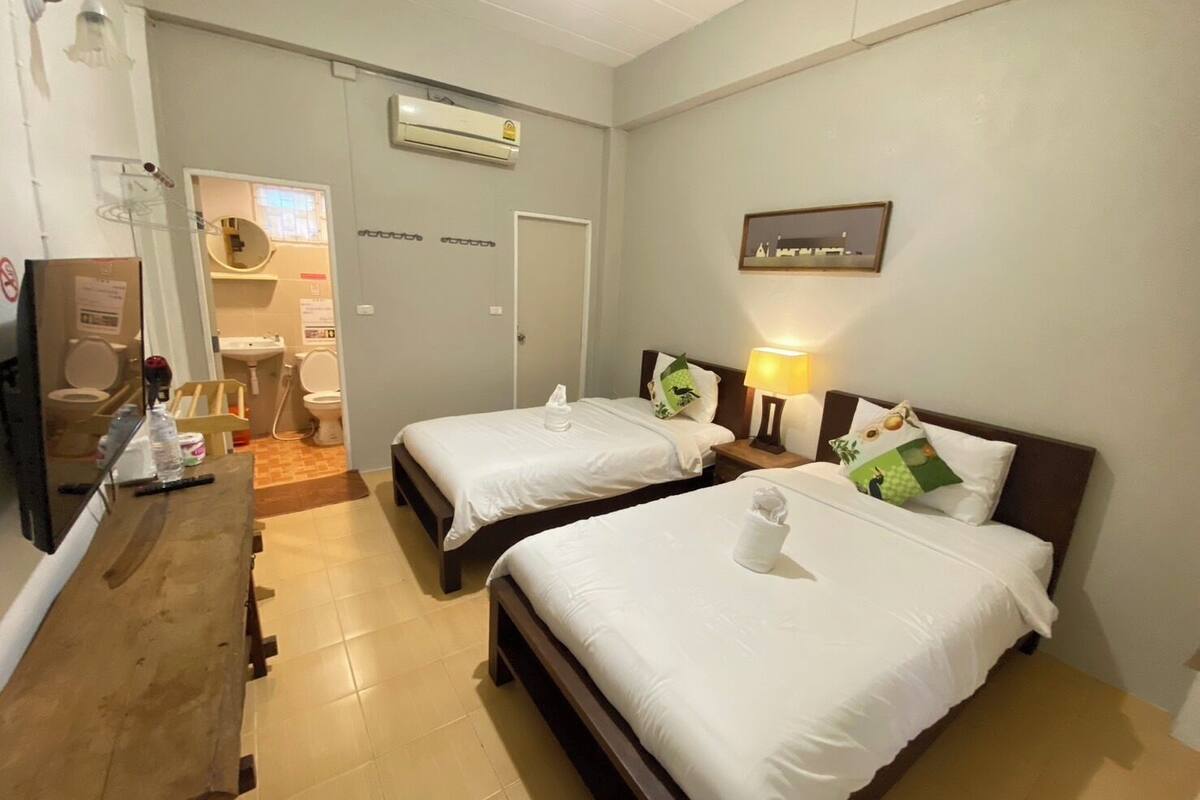 Une chambre d’hôtel avec deux lits, un bureau, un miroir, un climatiseur et une salle de bain visible à travers une porte ouverte.