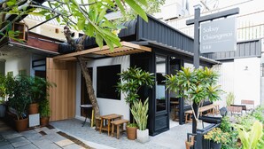 A modern outdoor seating area with wooden tables and chairs, surrounded by potted plants and a pergola.