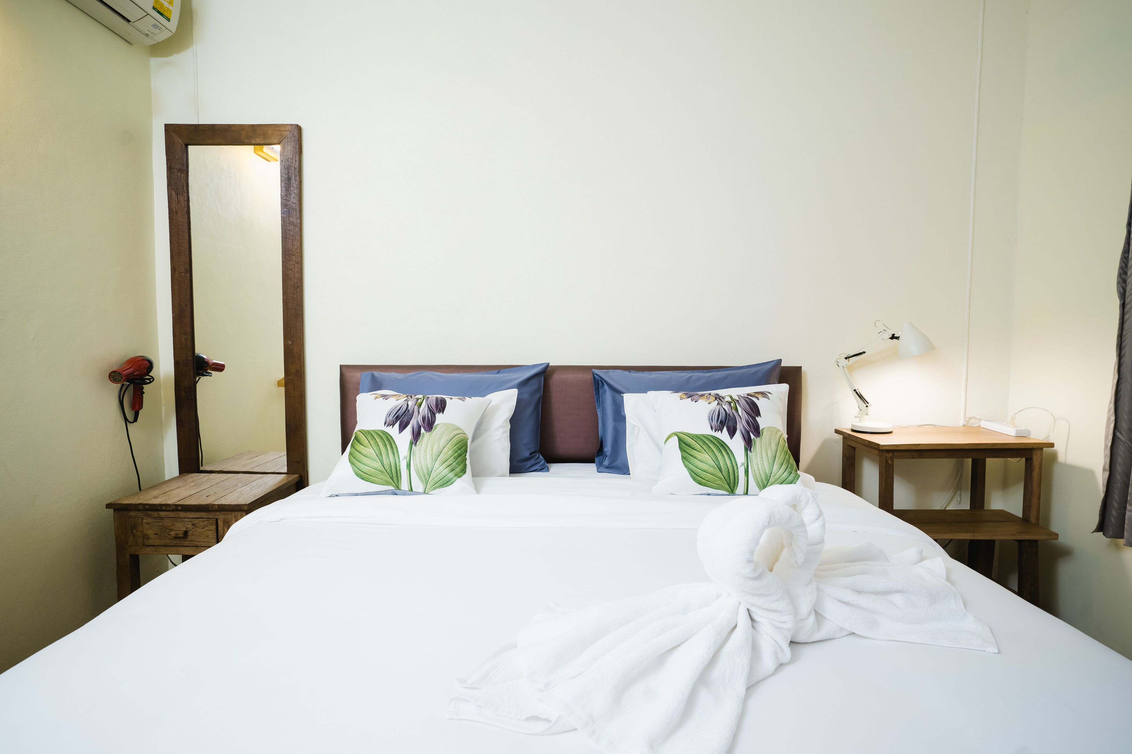 A neatly made bed with floral pillows, a wooden nightstand with a lamp, and a mirror on the wall.