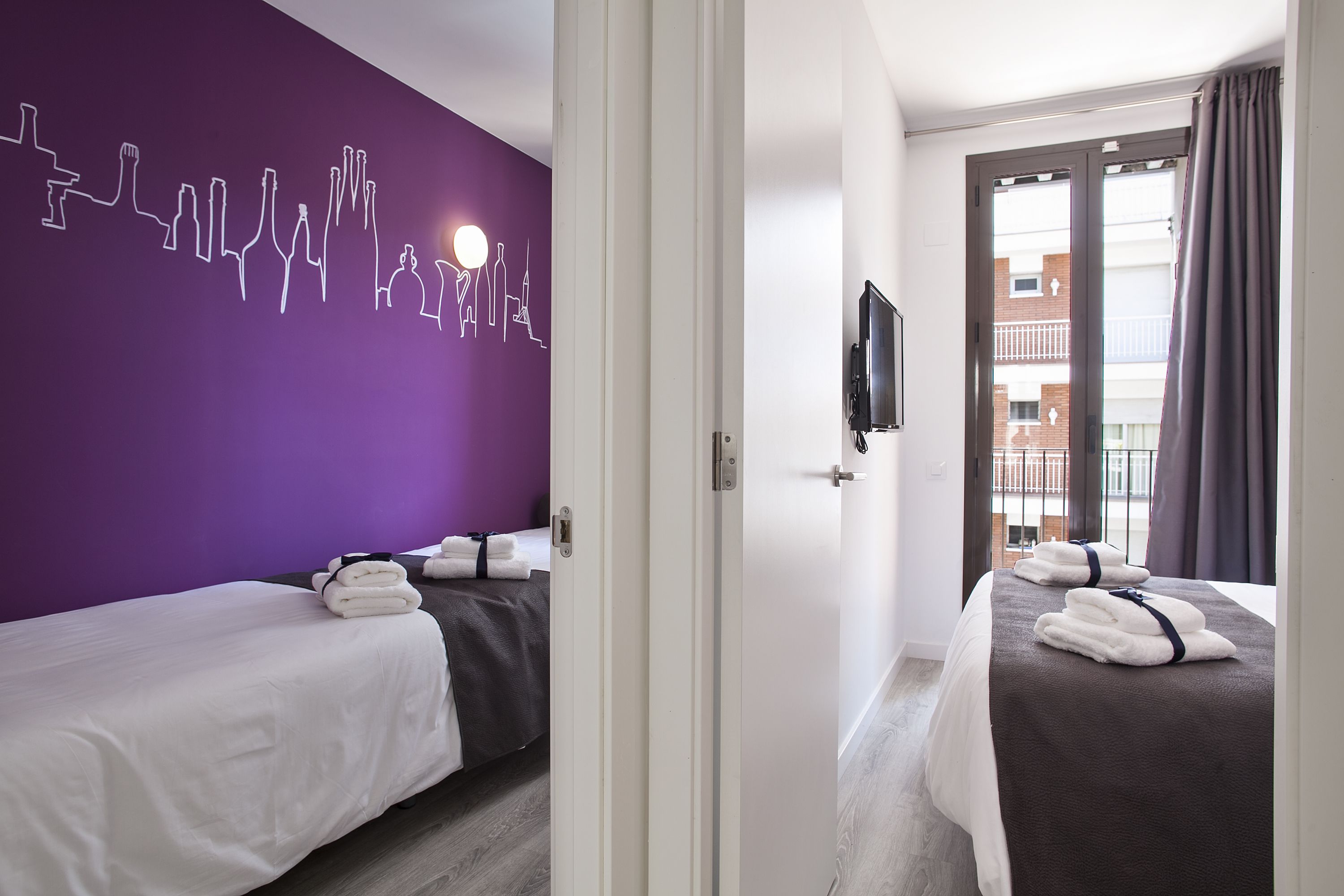 Une chambre d’hôtel avec deux lits, un mur violet orné d’un motif représentant le paysage urbain, un téléviseur fixé au mur et un balcon offrant une vue sur la ville.