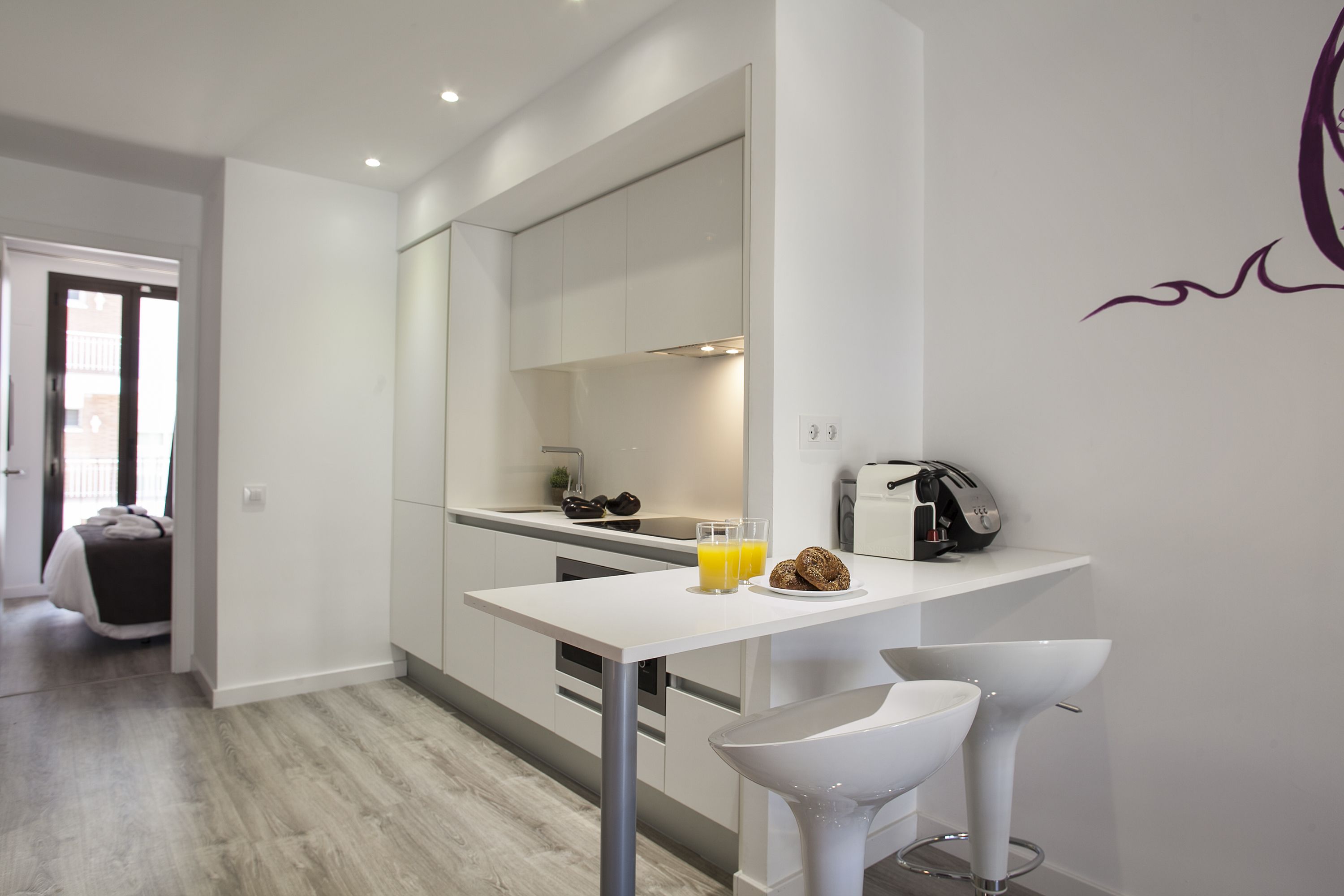 A modern kitchen with white cabinetry, a white countertop, and a breakfast bar with two bar stools. There is a toaster and a coffee machine on the countertop.