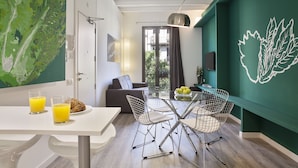 Uma sala de estar moderna com área de refeições, apresentando um sofá, uma mesa de vidro com cadeiras e uma grande parede verde com design de folhagens.