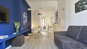 Uma sala moderna com um sofá cinzento, uma área de refeições com mesa e cadeiras, e uma TV fixada na parede azul.
