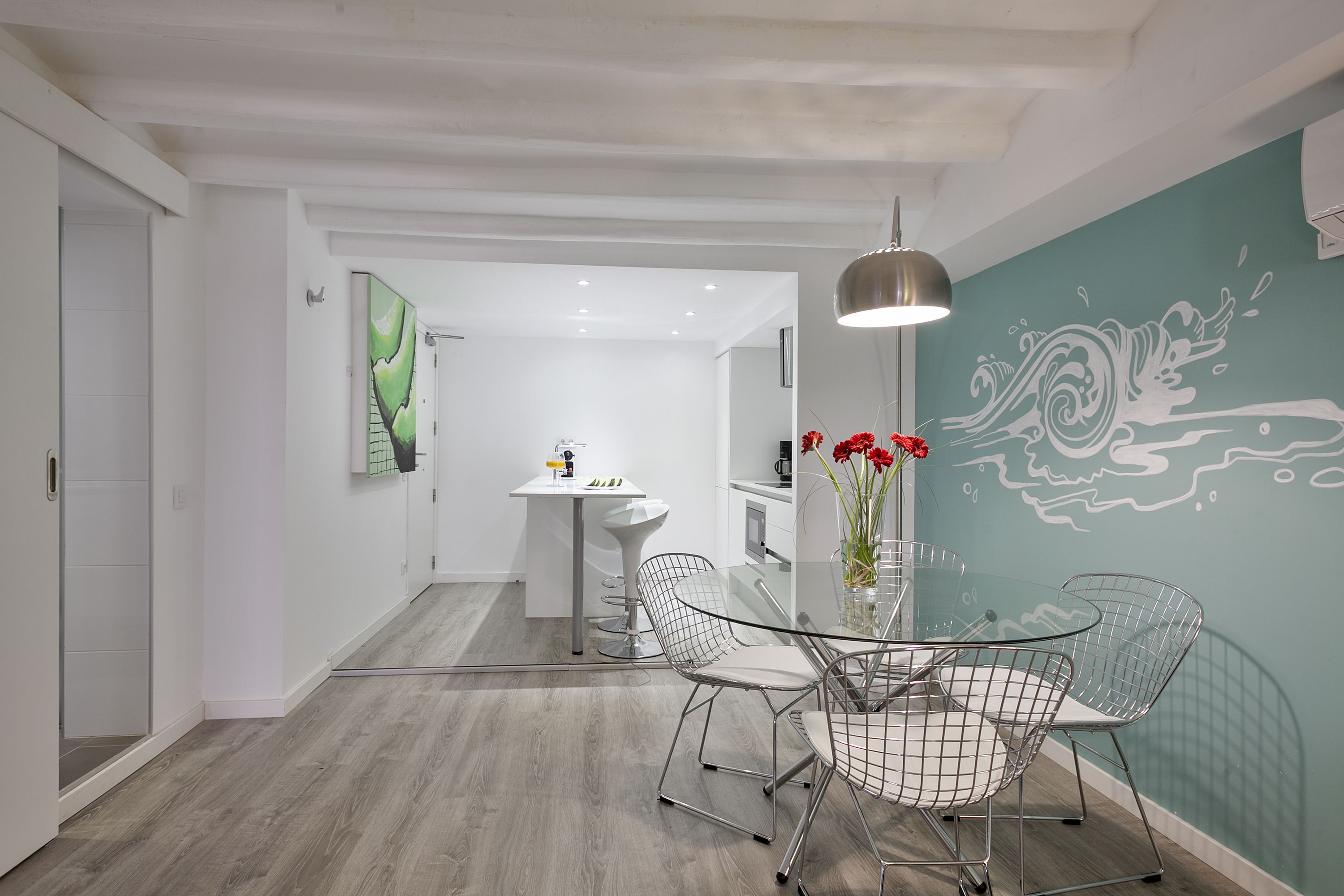 A modern kitchen with a glass dining table, wire chairs, and a pendant light.