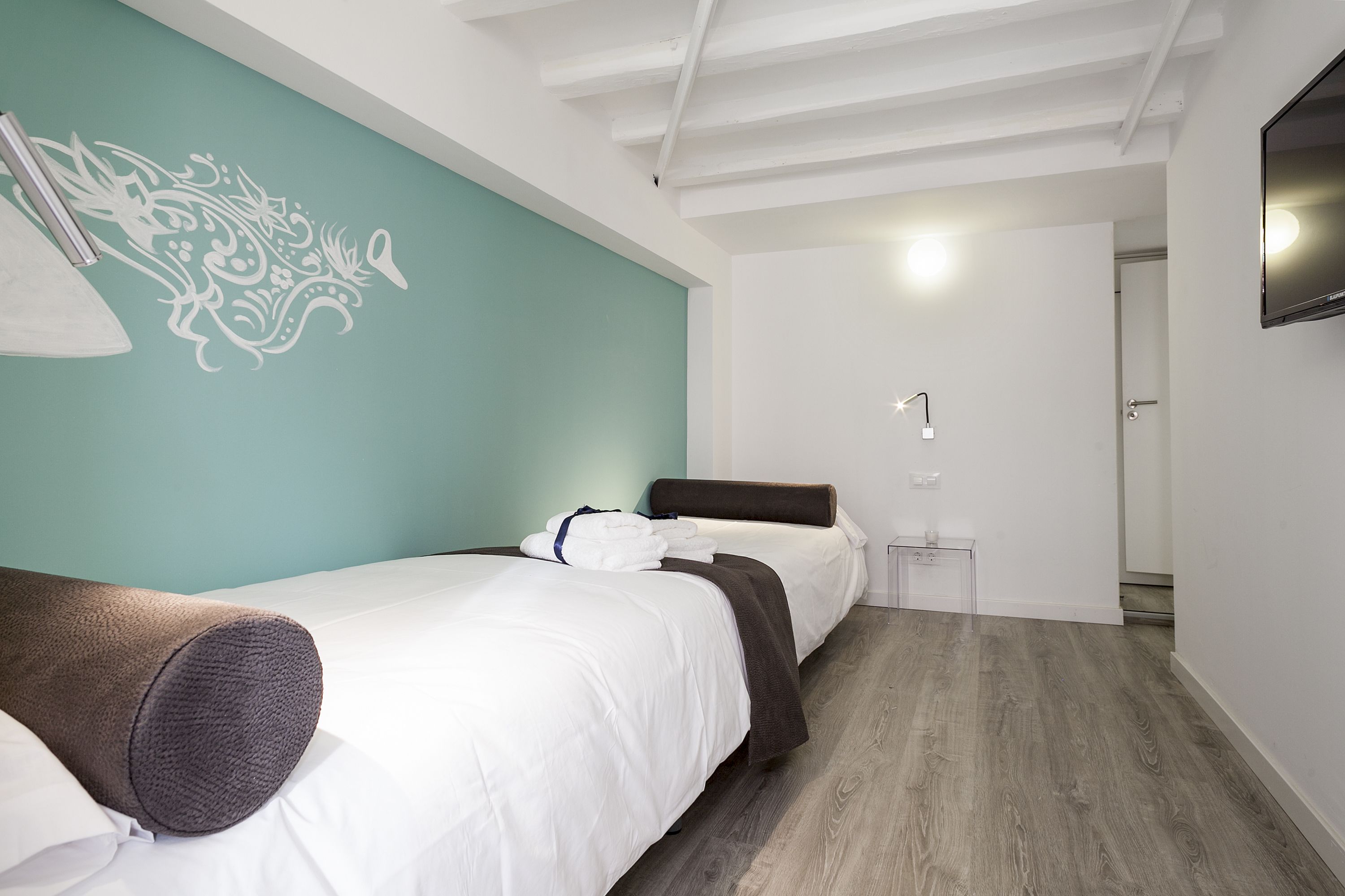 A modern hotel room with two beds, a teal accent wall, and a wooden floor.