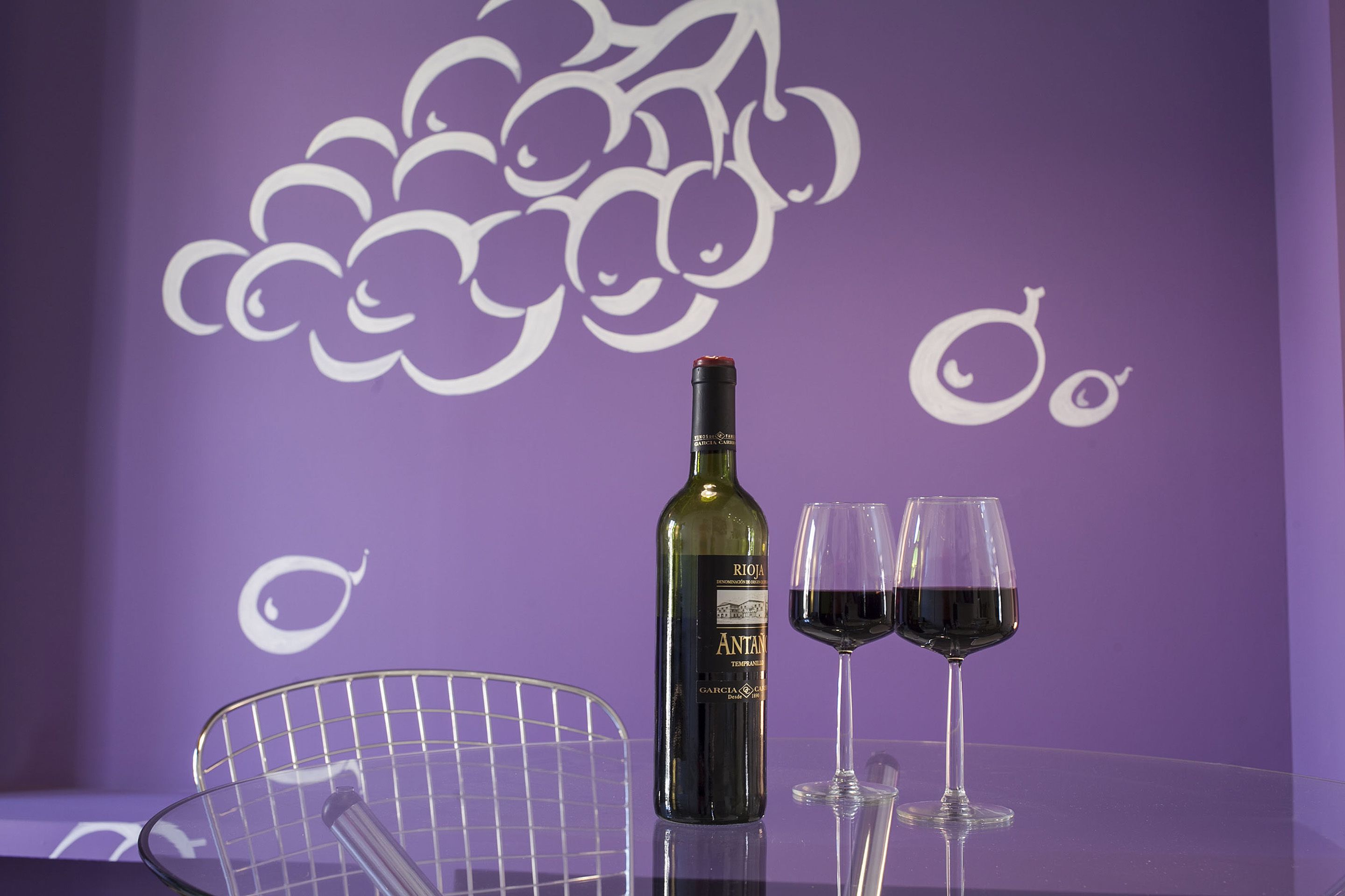 Una mesa con una botella de vino y dos copas, una raqueta de tenis, y una pared morada con diseño de parra.