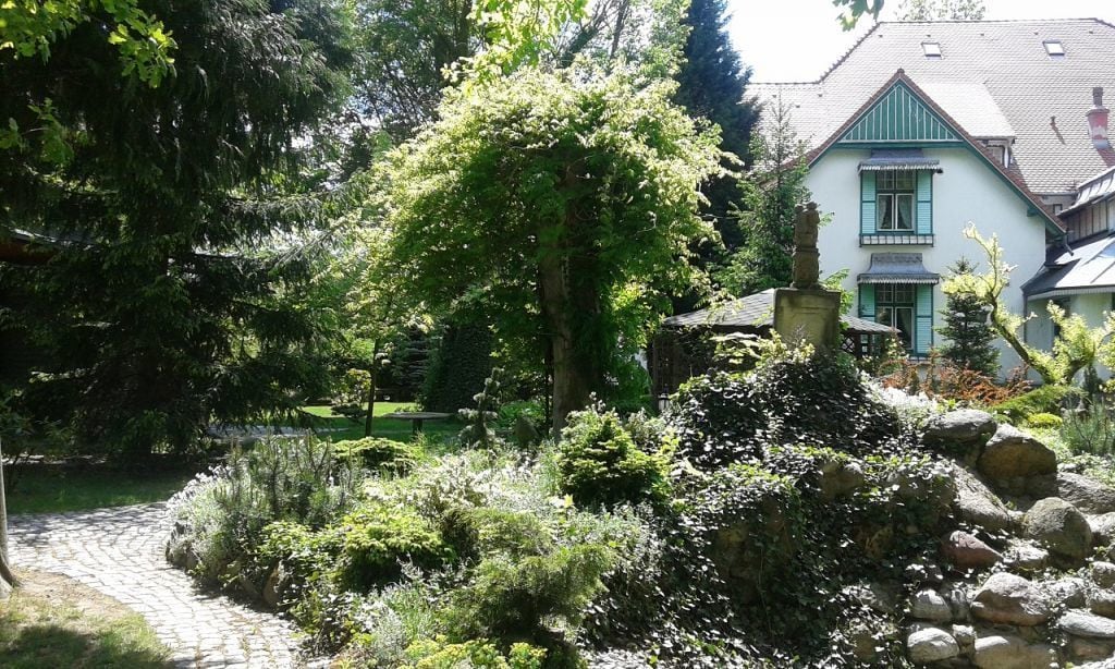 A garden with a stone pathway, a large tree, and a building with a pitched roof and shutters.