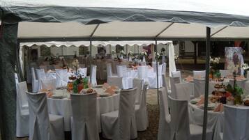 Een evenementenlocatie buiten, met ronde tafels met witte tafelkleden en stoelen, onder een grote tent.
