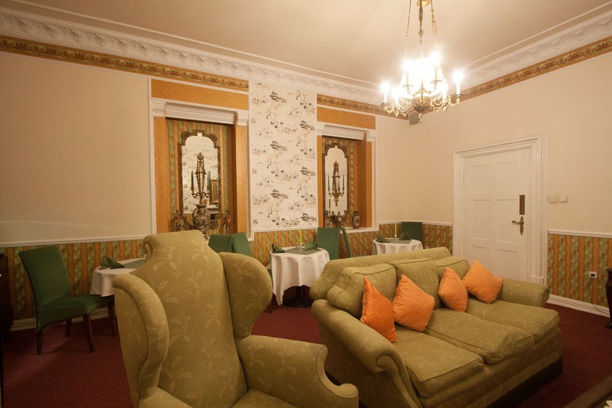 A room with a green sofa, matching armchairs, and a chandelier. There are two small tables with white tablecloths and green chairs. The walls have floral wallpaper, and there's a white door with a gold handle.