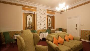A room with a green sofa, matching armchairs, and a chandelier. There are two small tables with white tablecloths and green chairs. The walls have floral wallpaper, and there's a white door with a gold handle.