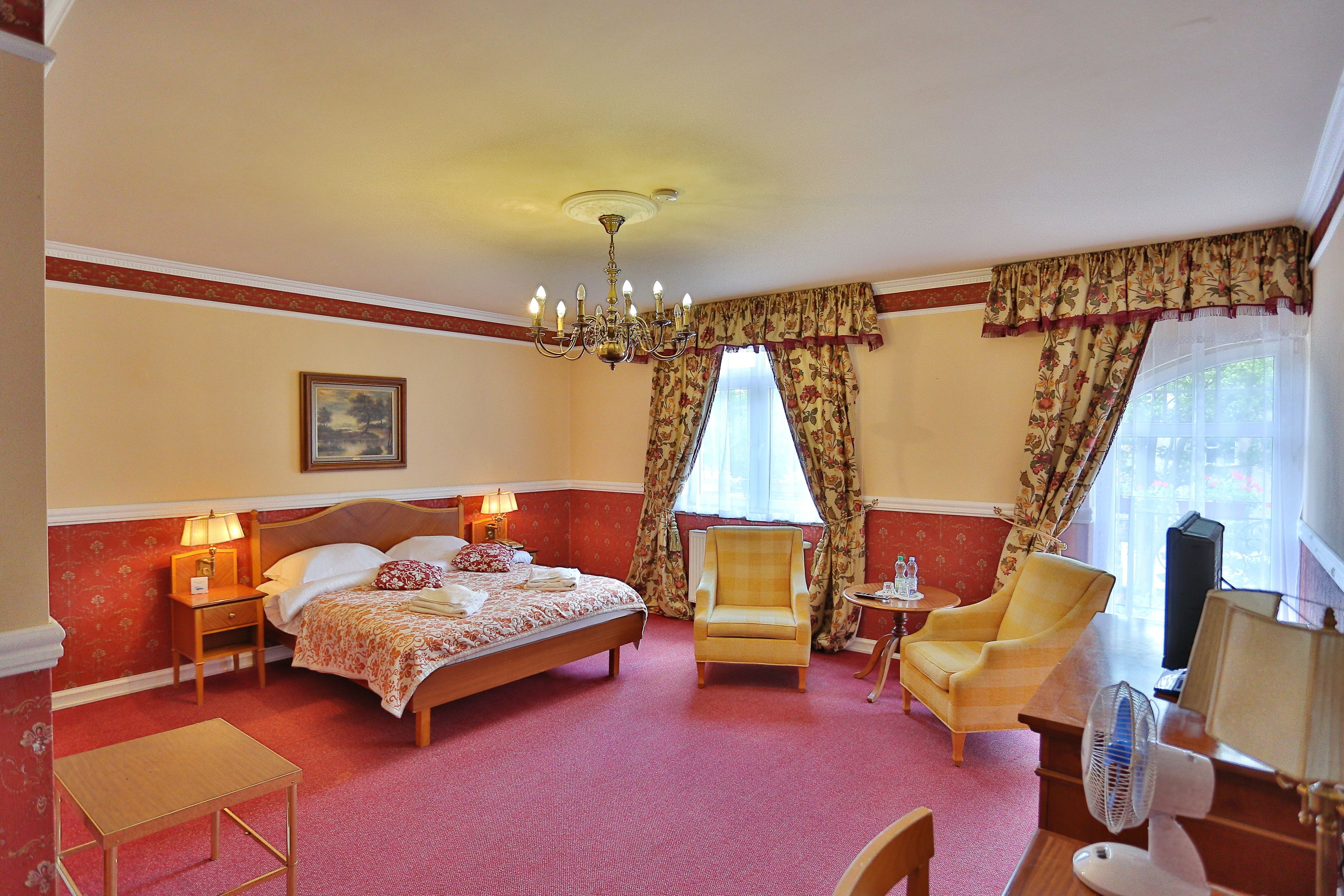 A hotel room with a bed, two armchairs, a desk with a fan, and a nightstand with a lamp.