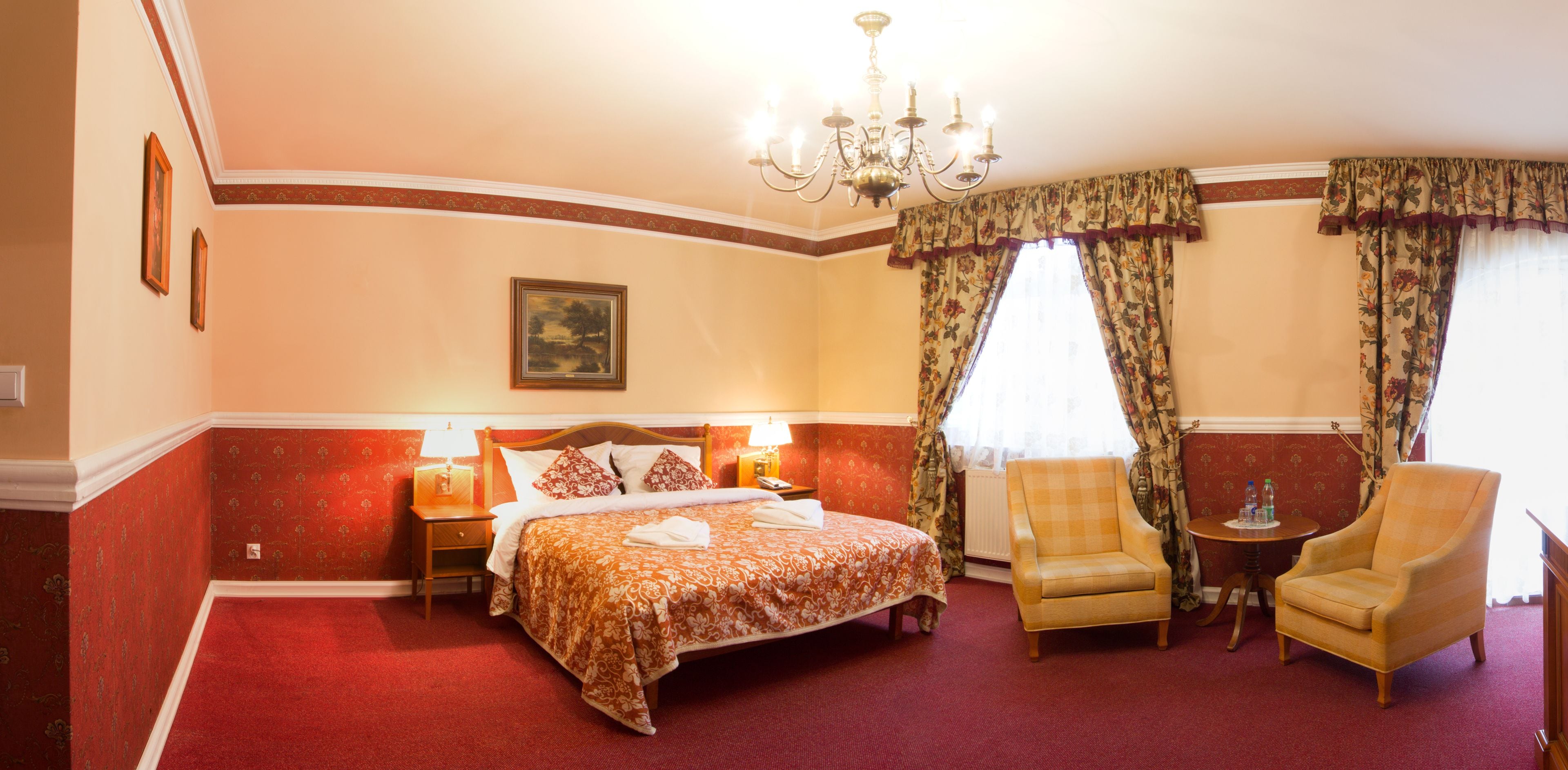 Habitación de hotel con una cama grande, dos sillones, una mesita, una araña y cortinas estampadas con motivos florales.
