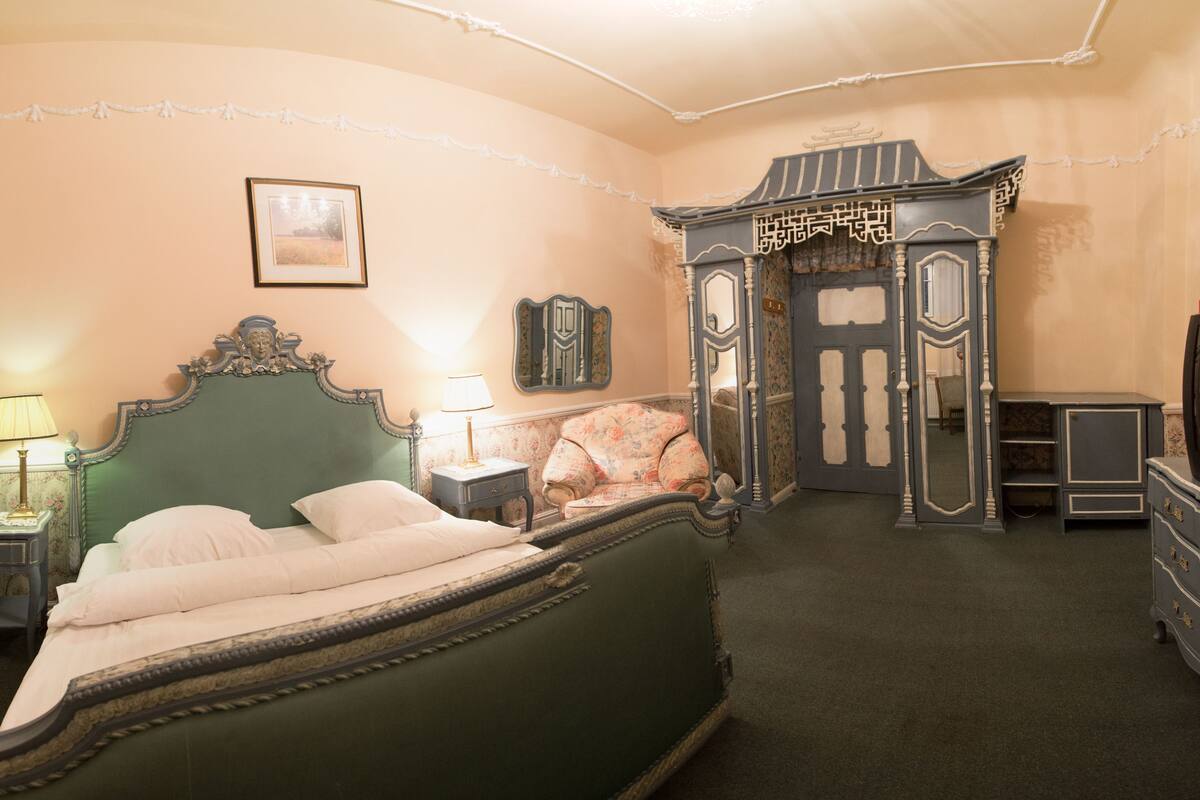 Ein traditionell eingerichtetes Schlafzimmer mit einem grünen, gepolsterten Bett, zwei Nachttischen mit Lampen, einem blumengemusterten Sessel und einem dekorativen Schrank.