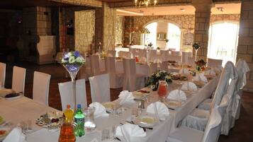 Een balzaal met meerdere ronde tafels gedekt voor een formeel evenement, stenen muren en een kroonluchter.
