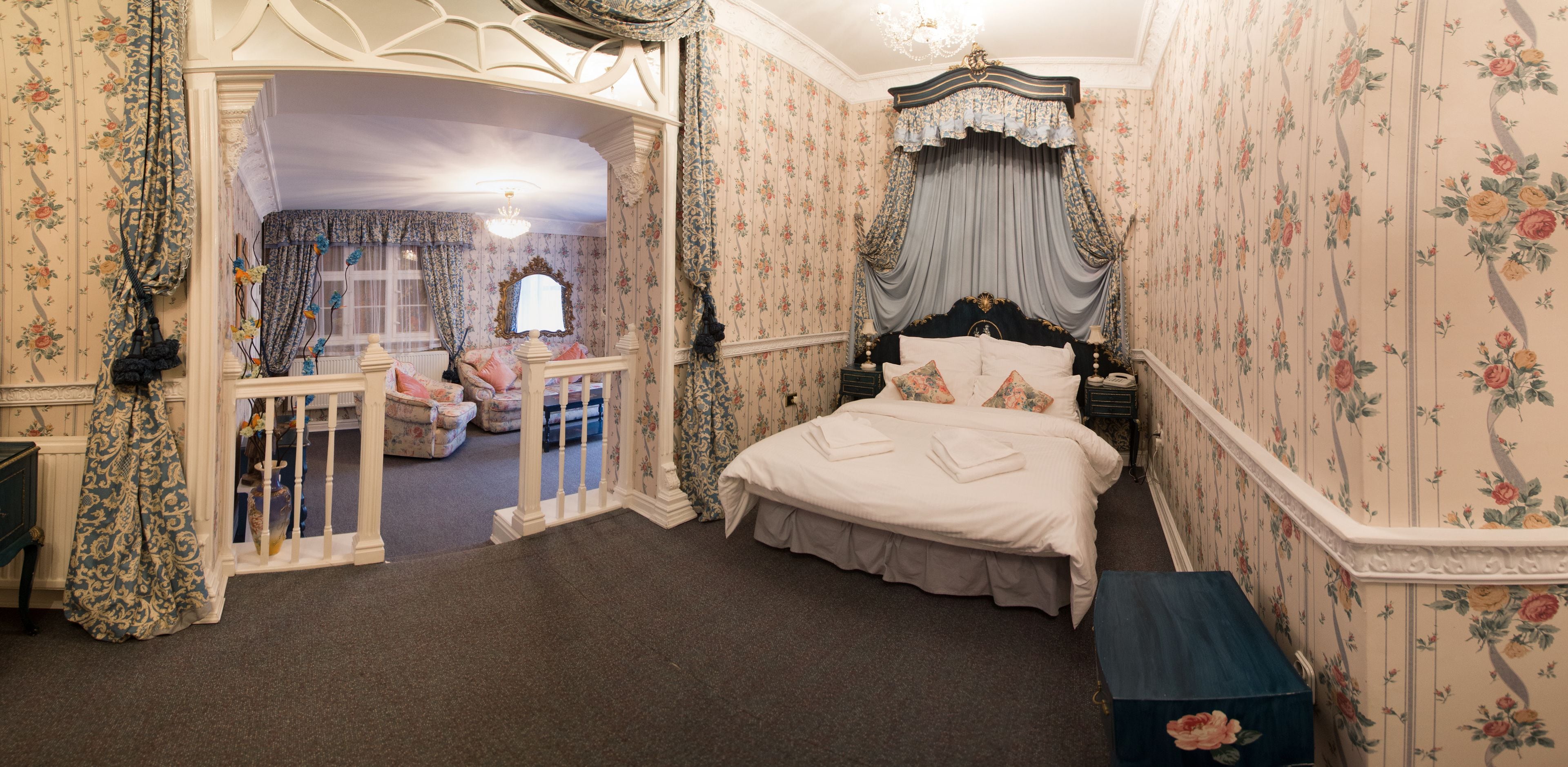 Slaapkamer met een groot bed, een hemelbed, bloemenbehang en uitzicht op een andere kamer door een geopende deur.