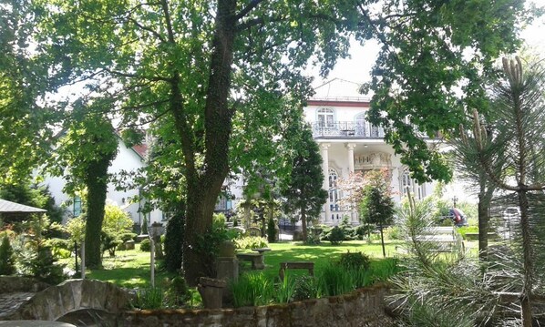 A garden with a stone bridge, lush greenery, and a building with a balcony in the background.