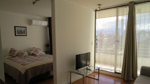 Een hotelkamer met een bed, een televisie op een glazen tafeltje, een balkon met gordijnen en uitzicht op de stad.