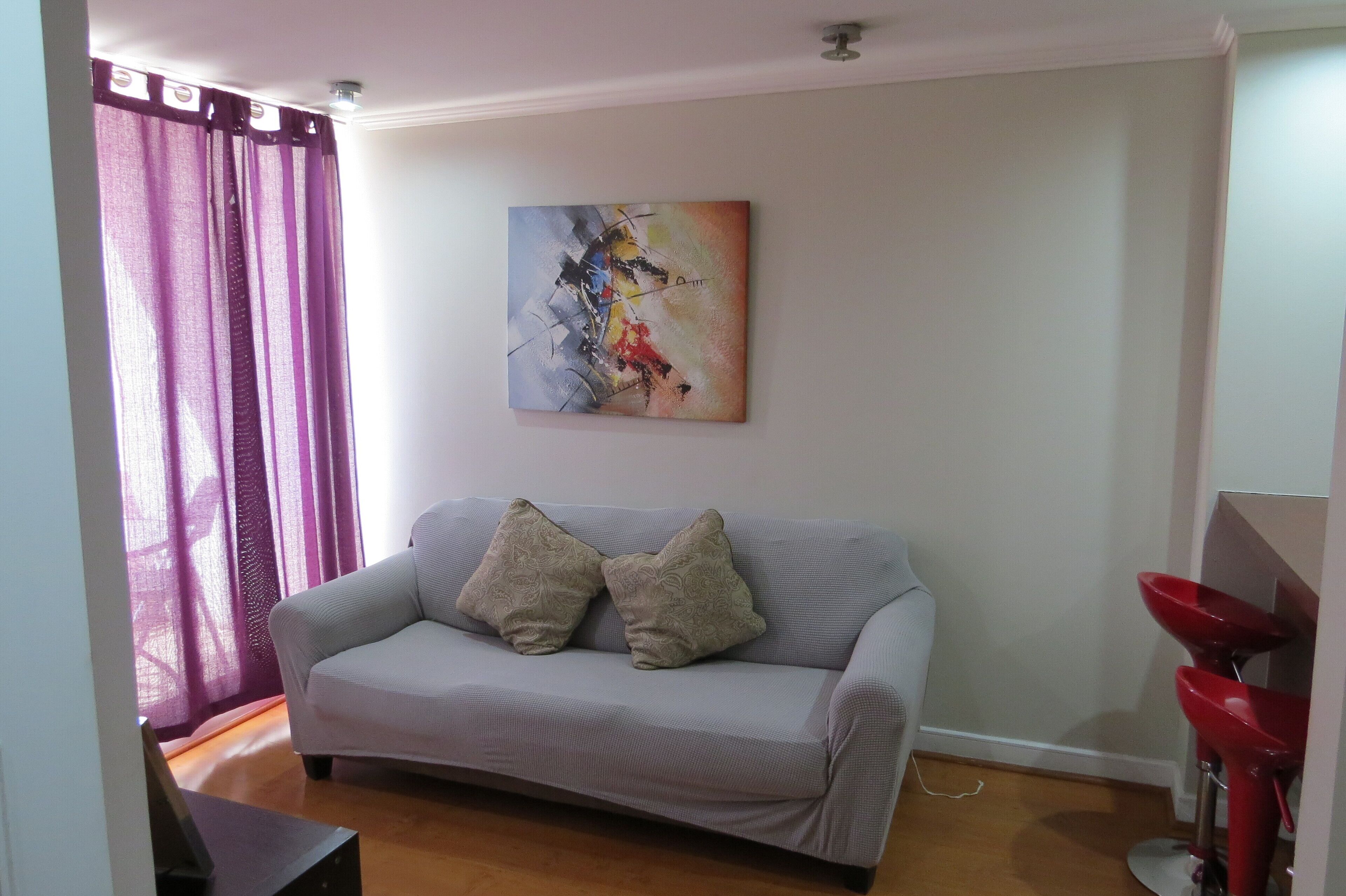 A living room with a grey sofa, two patterned cushions, a colorful abstract painting on the wall, and a red sink in the corner.