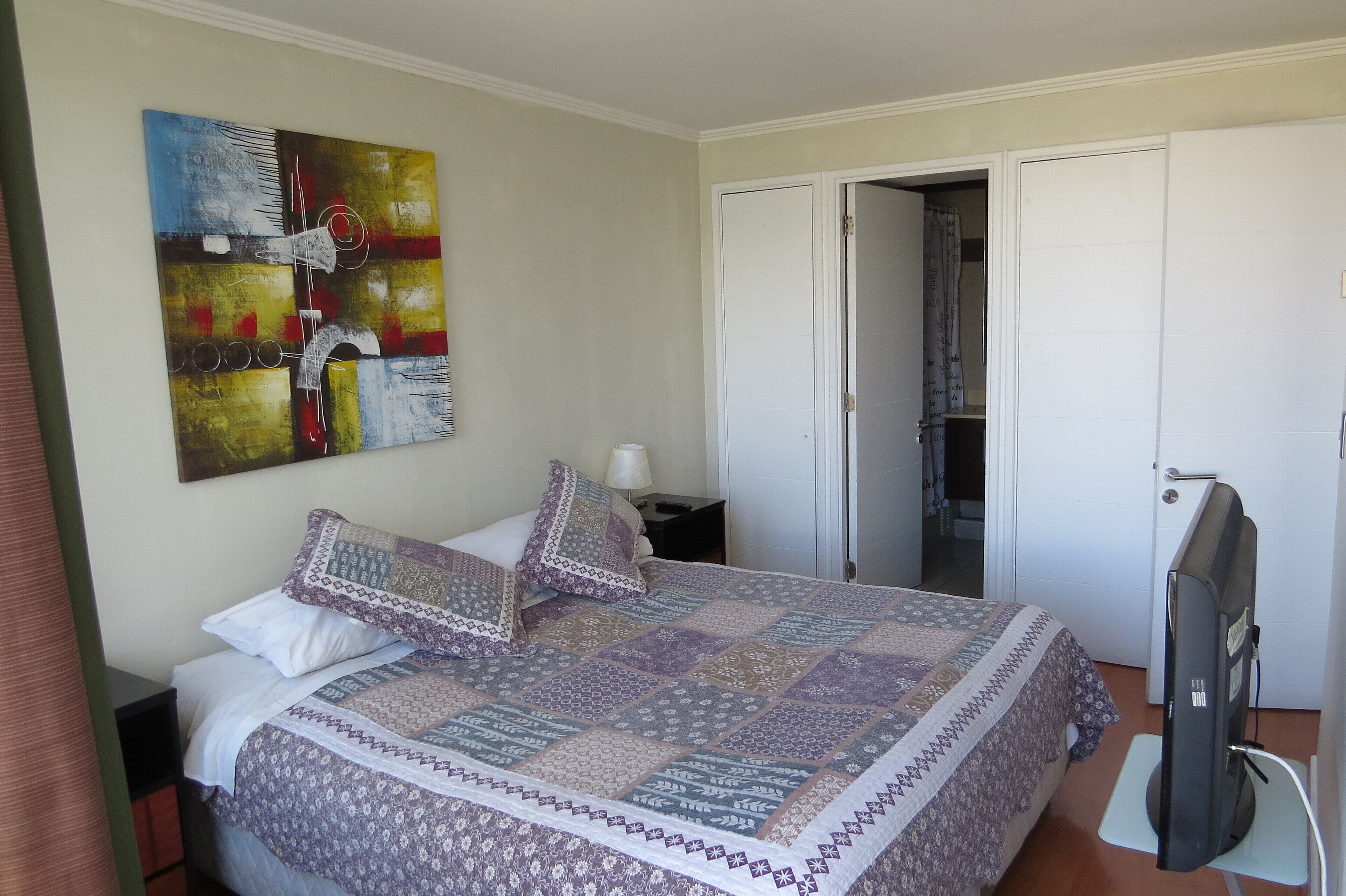 A neatly made bed with a patterned quilt, a wall art piece, a television, and a wardrobe.