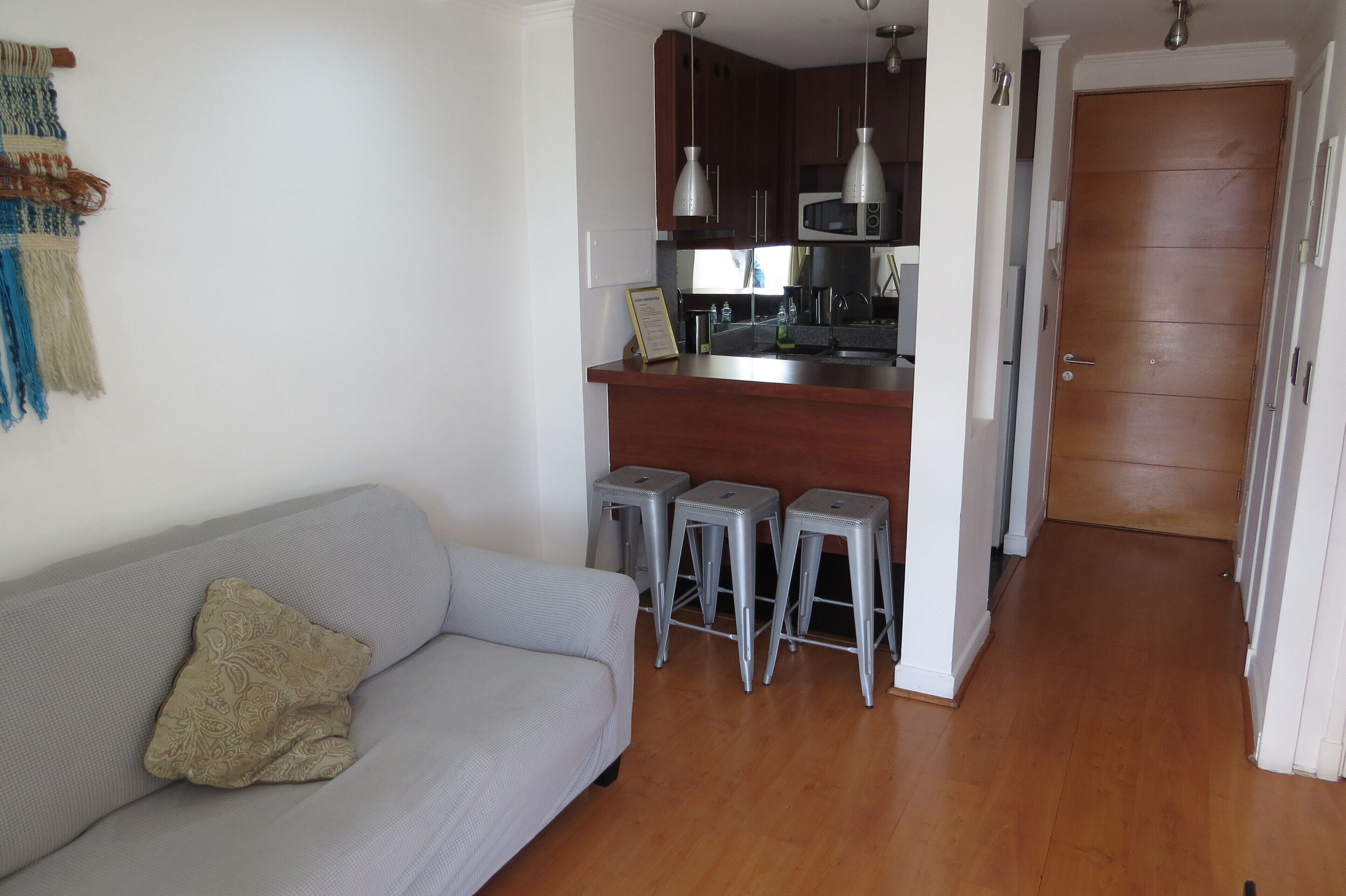 A living room with a grey sofa and a patterned pillow, a kitchen with a microwave and hanging lights, and a wooden door.