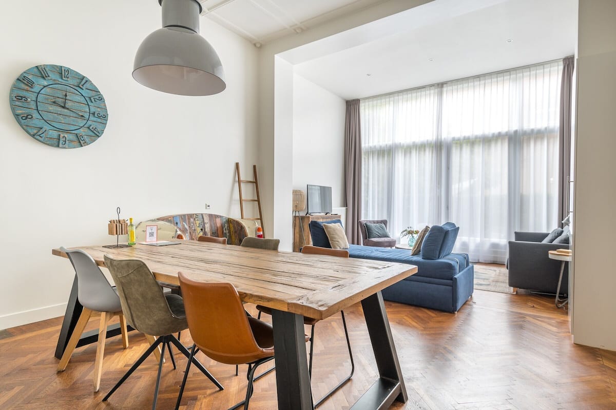Een moderne woonkamer met een houten eettafel, grijze en oranje stoelen, een blauwe bank en een groot raam met dunne gordijnen.