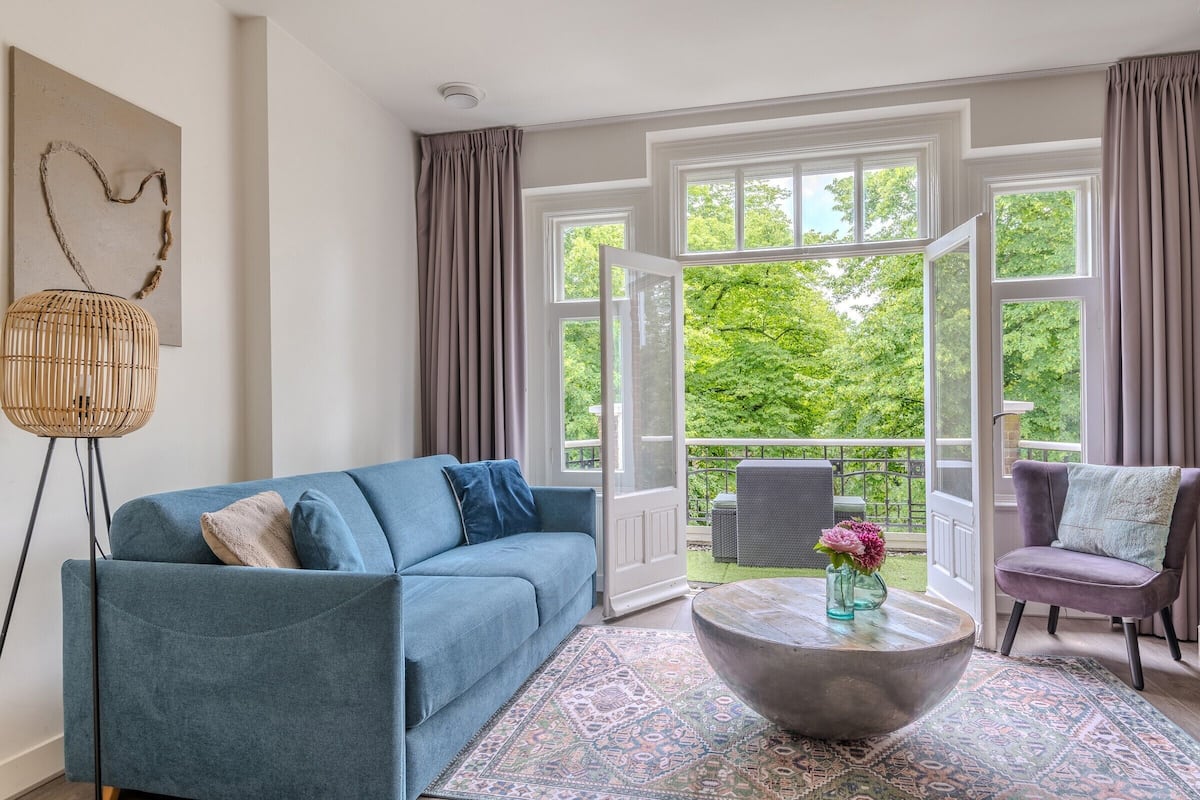 Een moderne woonkamer met een blauwe bank, een ronde salontafel met een vaas, en uitzicht op een balkon met beplanting.