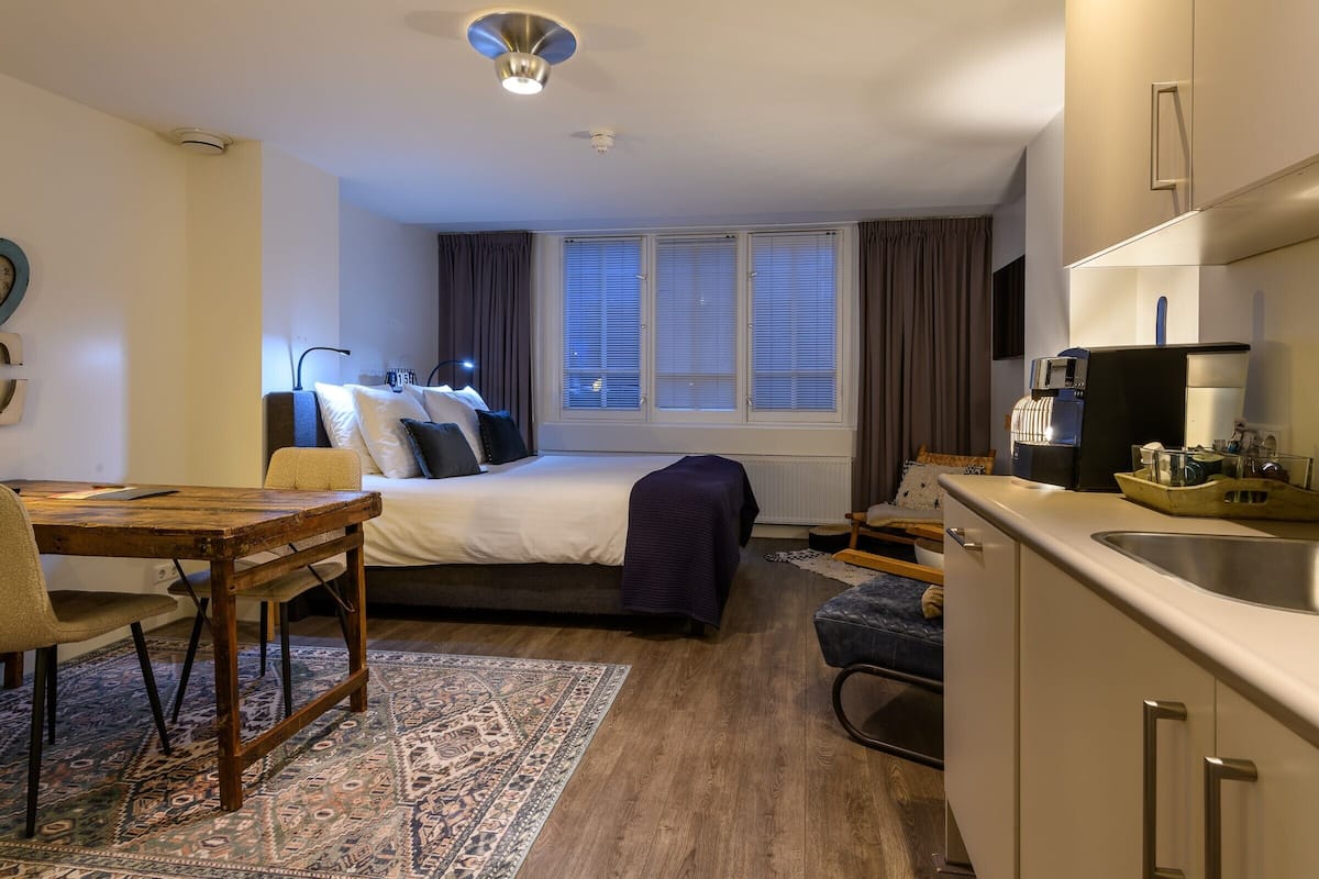 Een compacte hotelkamer met een bed, een klein eettafeltje, een kitchenette en een groot raam met gordijnen.