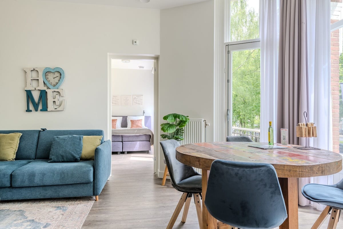 Een moderne woonkamer met een blauwe bank, een houten eettafel met stoelen, en uitzicht op een tuin door grote ramen.