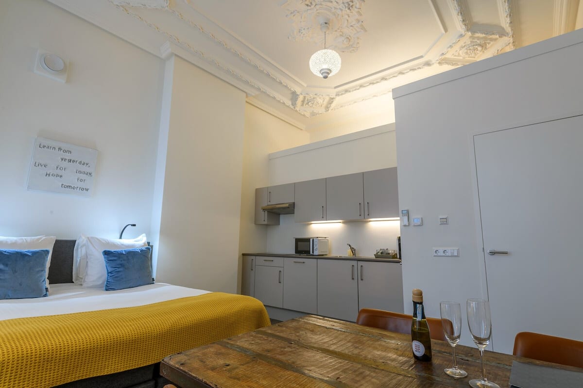 Een compacte hotelkamer met een bed, een houten tafel met een fles en glazen, een kitchenette en een decoratieve plafondlamp.