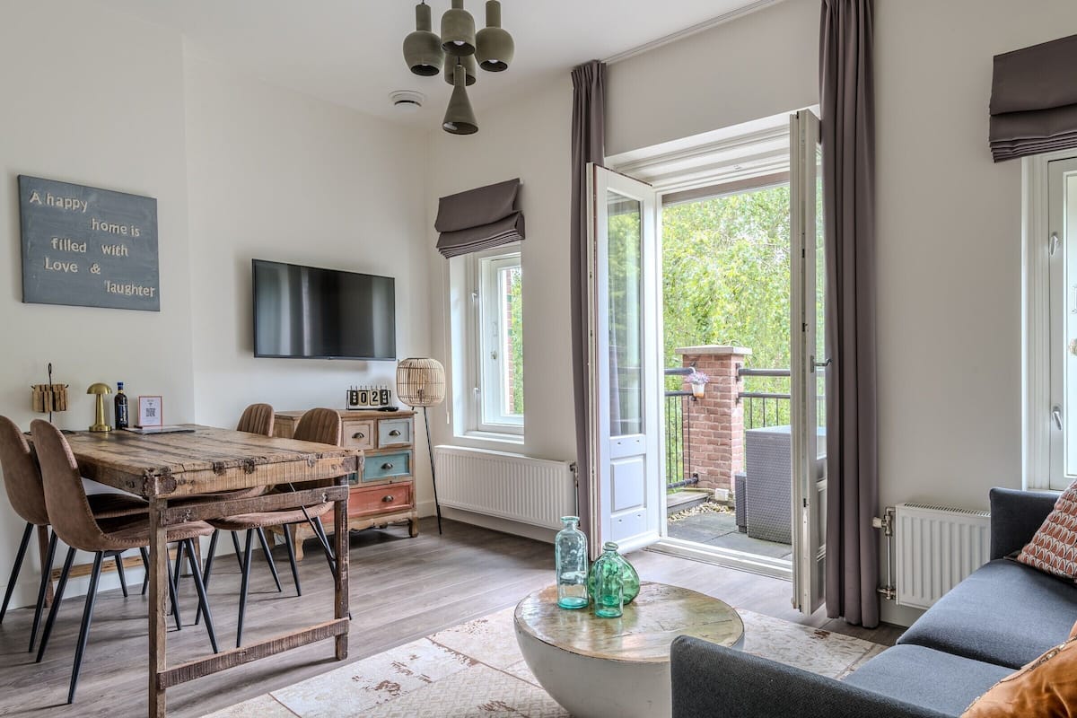 Een gezellige woonkamer met een houten eettafel, comfortabele zithoek, een flatscreen televisie en een groot raam met gordijnen.