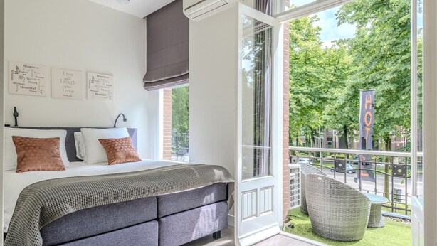 A bedroom with a large bed, a view of the outdoors, and a balcony with outdoor furniture.