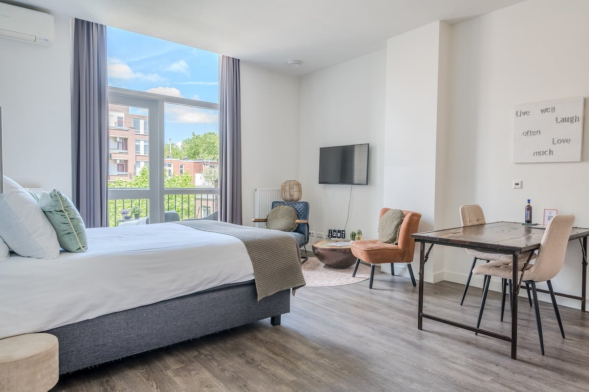 Een moderne hotelkamer met een groot bed, een eettafel met stoelen, een aan de muur bevestigde televisie en een balkon met uitzicht op gebouwen en bomen.