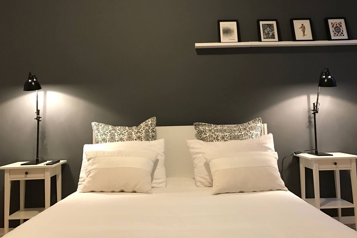 A neatly made bed with white linens and patterned pillows, flanked by two bedside tables with lamps, against a dark wall with a shelf above displaying framed pictures.
