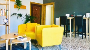 Una habitación con un sillón amarillo, una pequeña mesa redonda, una estantería, una planta en maceta, una puerta de madera y una zona de comedor con sillas y una mesa.