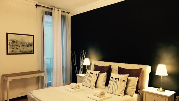 Una cama bien hecha con edredón blanco y almohadones, una mesita de noche con lámpara, una mesita auxiliar y un cuadro en la pared.