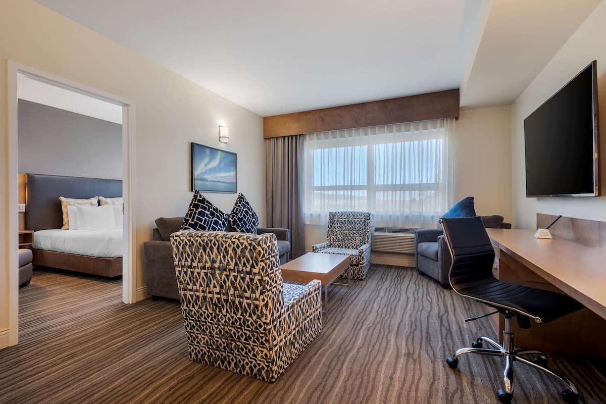 A hotel room with a bed, a sofa, a patterned armchair, a desk with a chair, a flat-screen TV, and a large window with curtains.