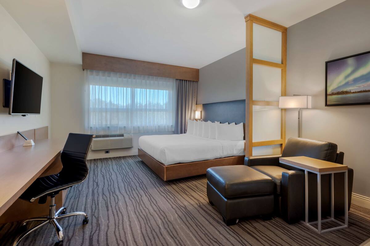 A hotel room with a bed, desk, chair, ottoman, and a TV.