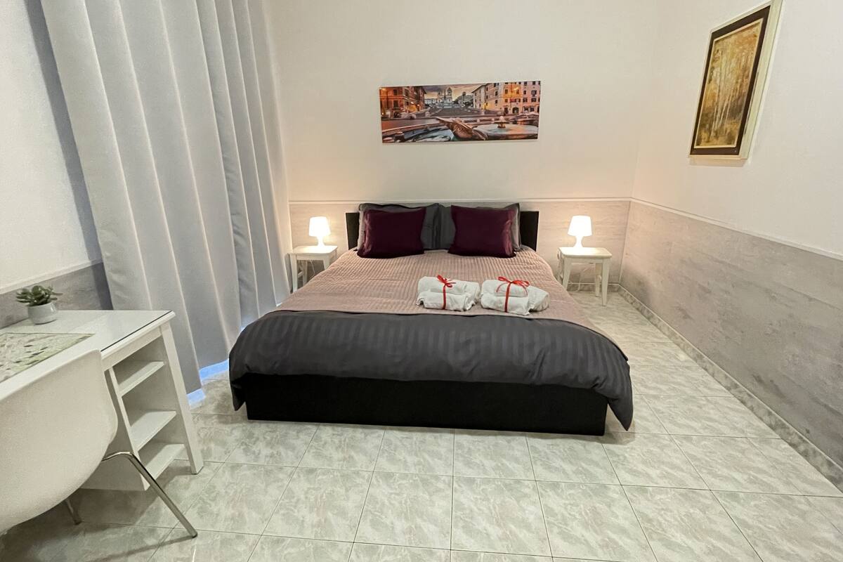 Quarto de hotel com cama, duas mesas de cabeceira com luminárias, escrivaninha, cadeira e um quadro na parede.