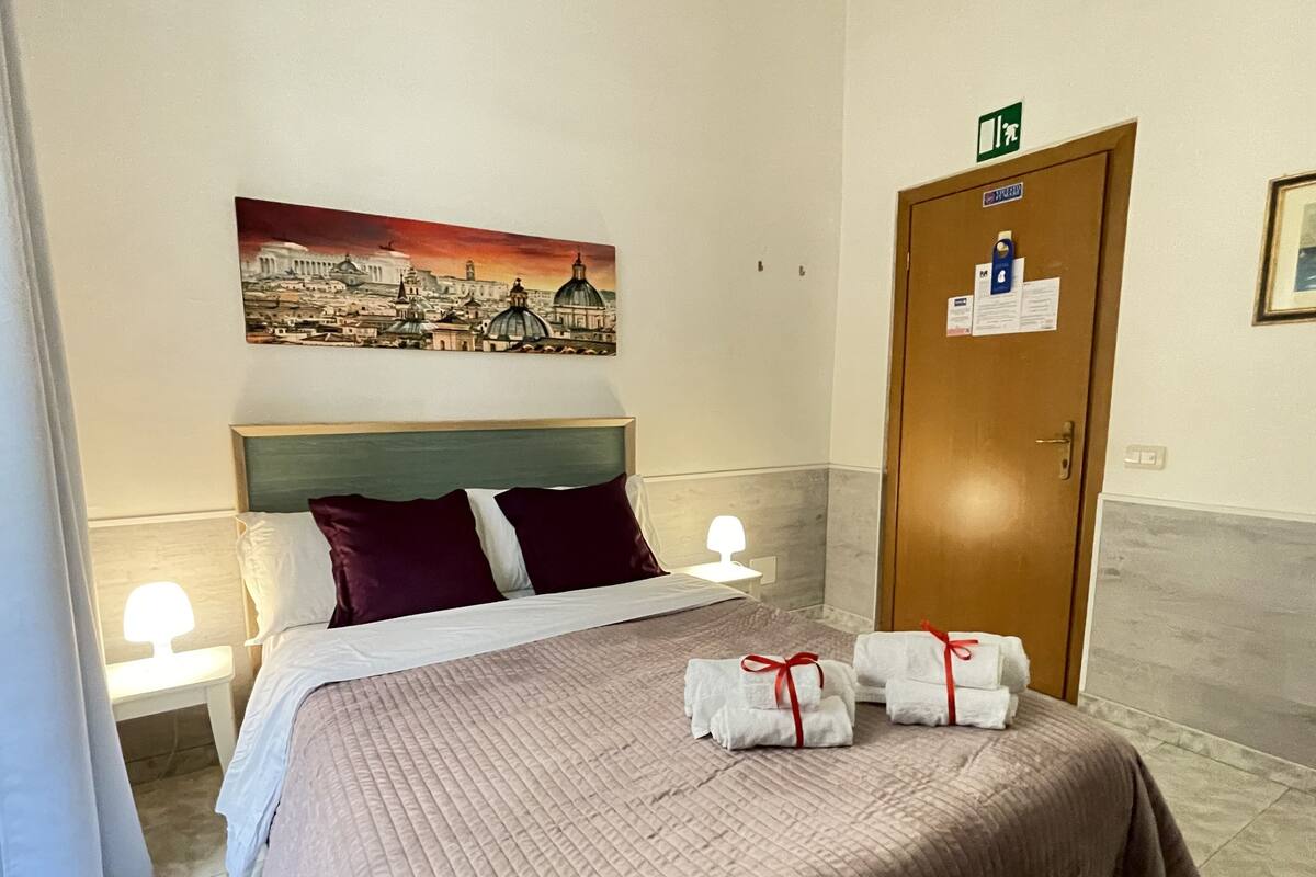 Quarto de hotel com uma cama, um quadro de uma paisagem urbana, uma porta com uma placa e toalhas sobre a cama.
