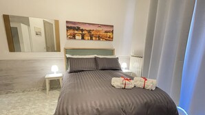 A neatly made bed with a dark brown bedspread and pillows, two bedside tables with lamps, a large framed picture of a cityscape, and a mirror on the wall.