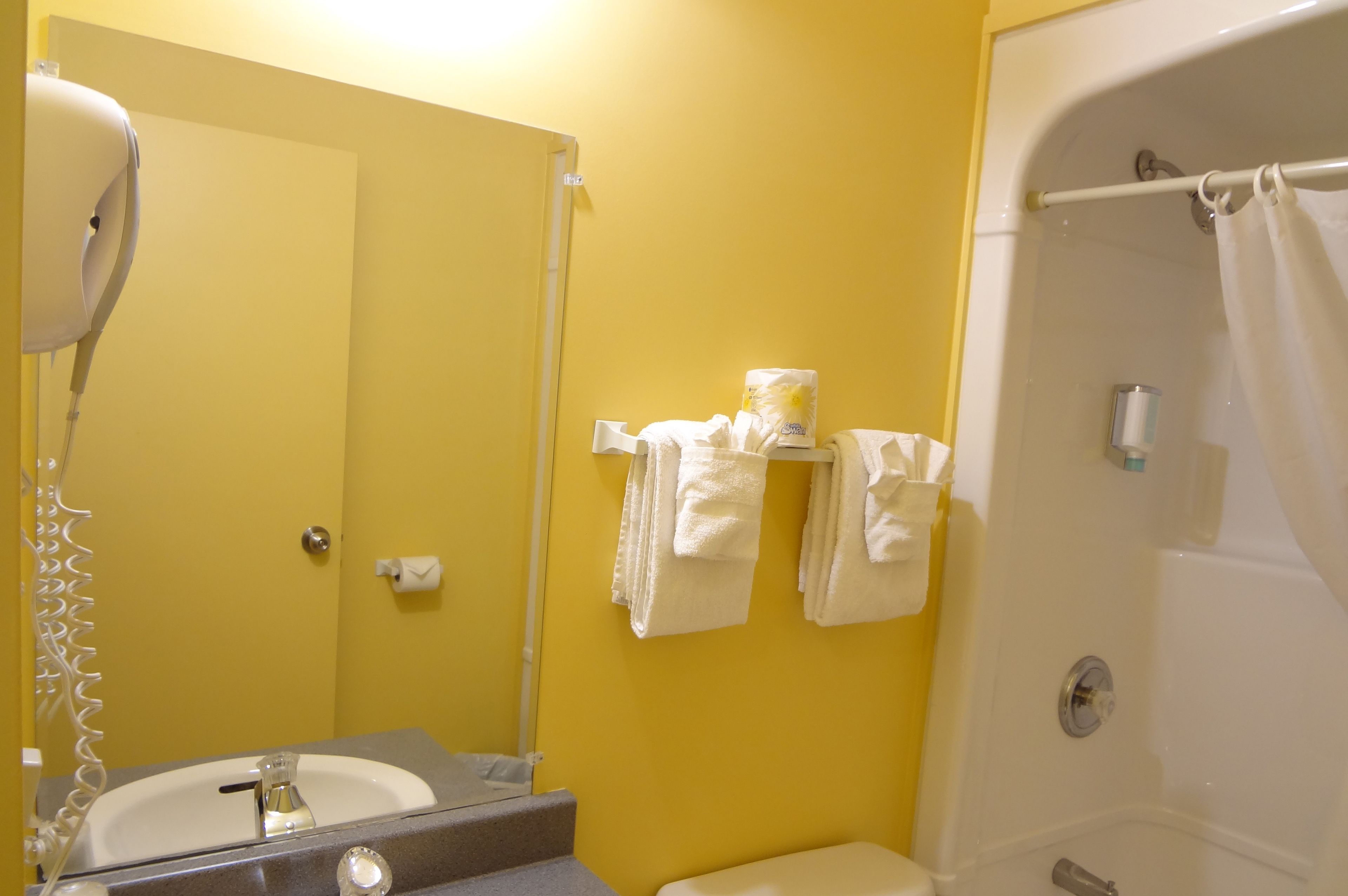 A bathroom with a yellow wall, a sink, a mirror, a hairdryer, a towel rack with towels, and a bathtub with a shower curtain.