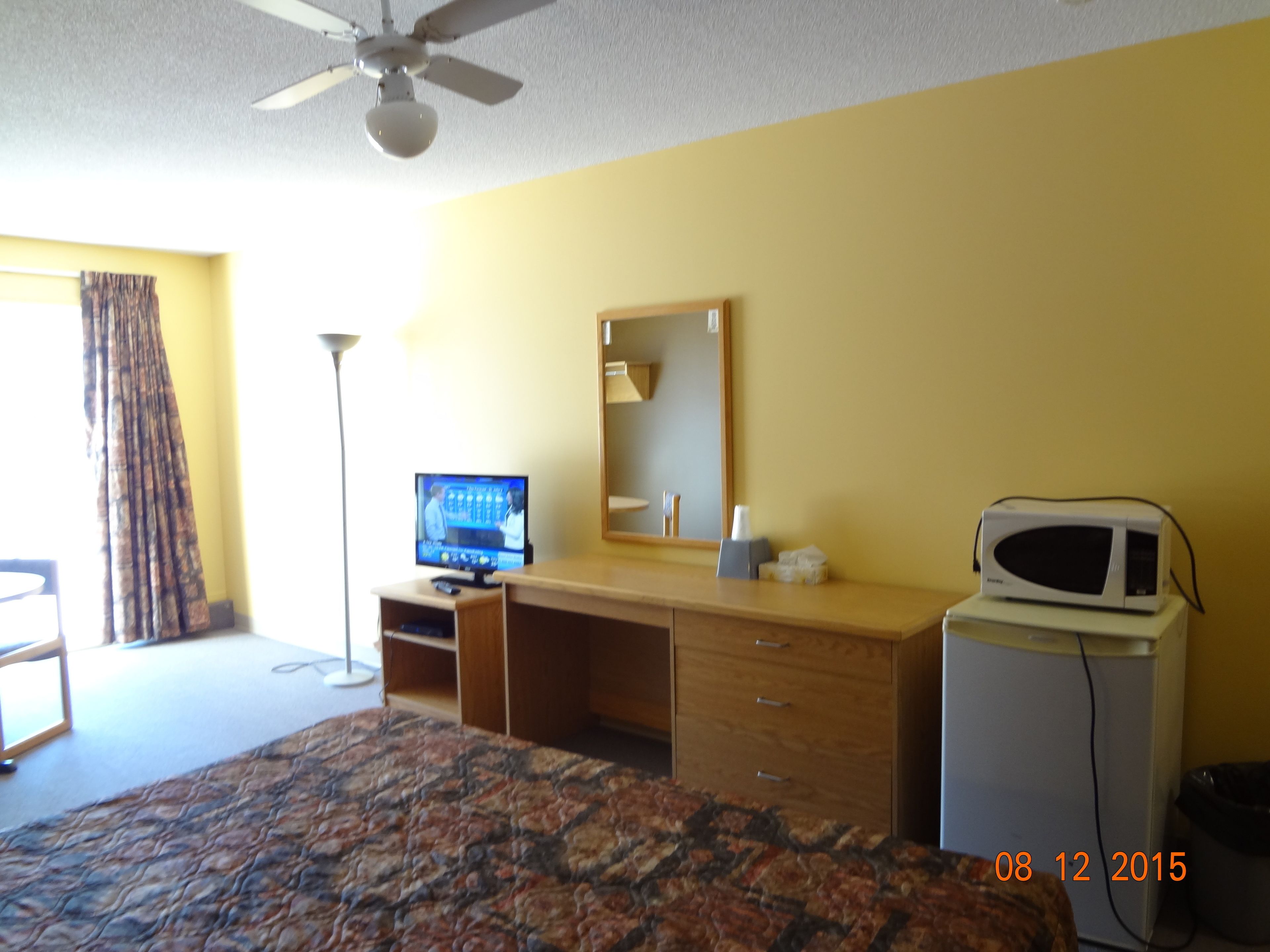 A hotel room with a bed, a desk, a television, a microwave, a mirror, and a window with curtains.