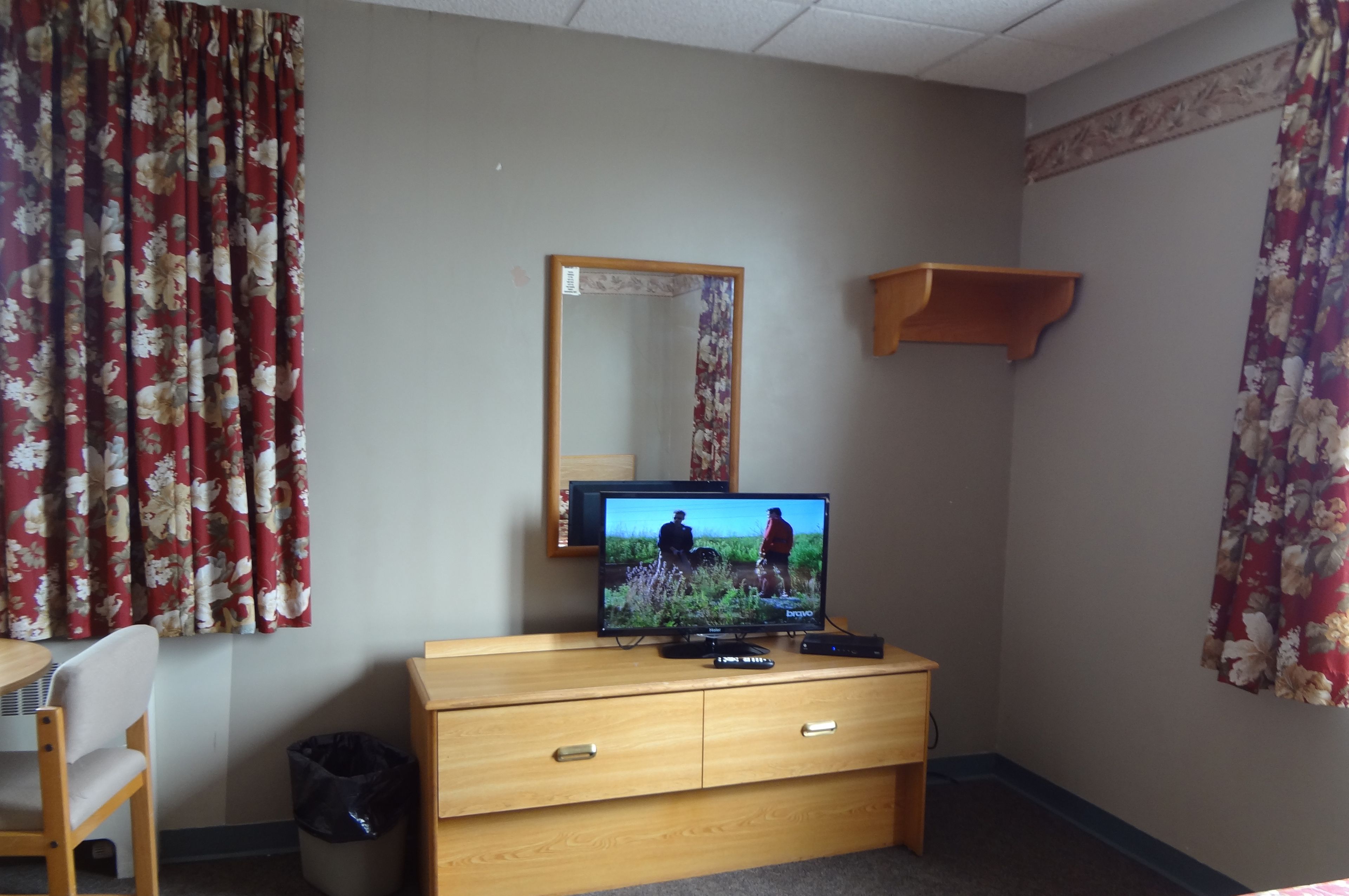 A hotel room with a wooden dresser, a television on top, a mirror above it, and floral curtains.