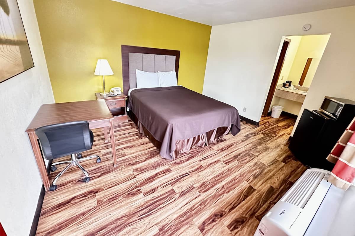 A hotel room with a bed, desk, chair, bedside table, lamp, and a small kitchenette.