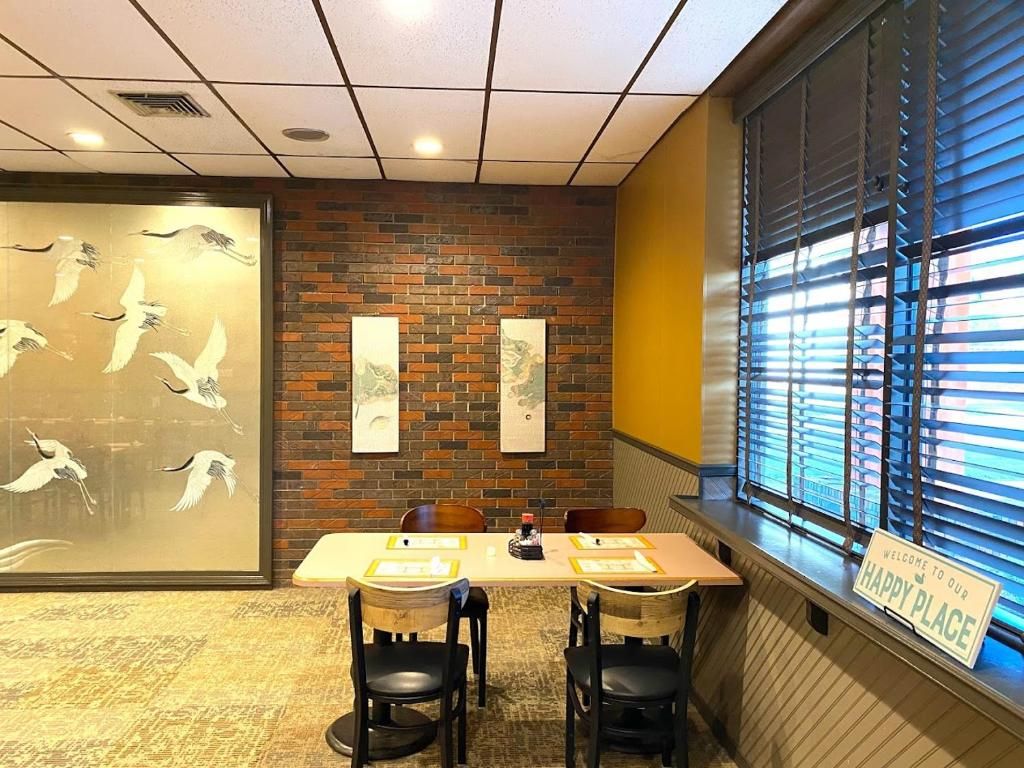 Una habitación con pared de ladrillo, un ventanal con persianas, una mesa con sillas y un letrero de bienvenida.