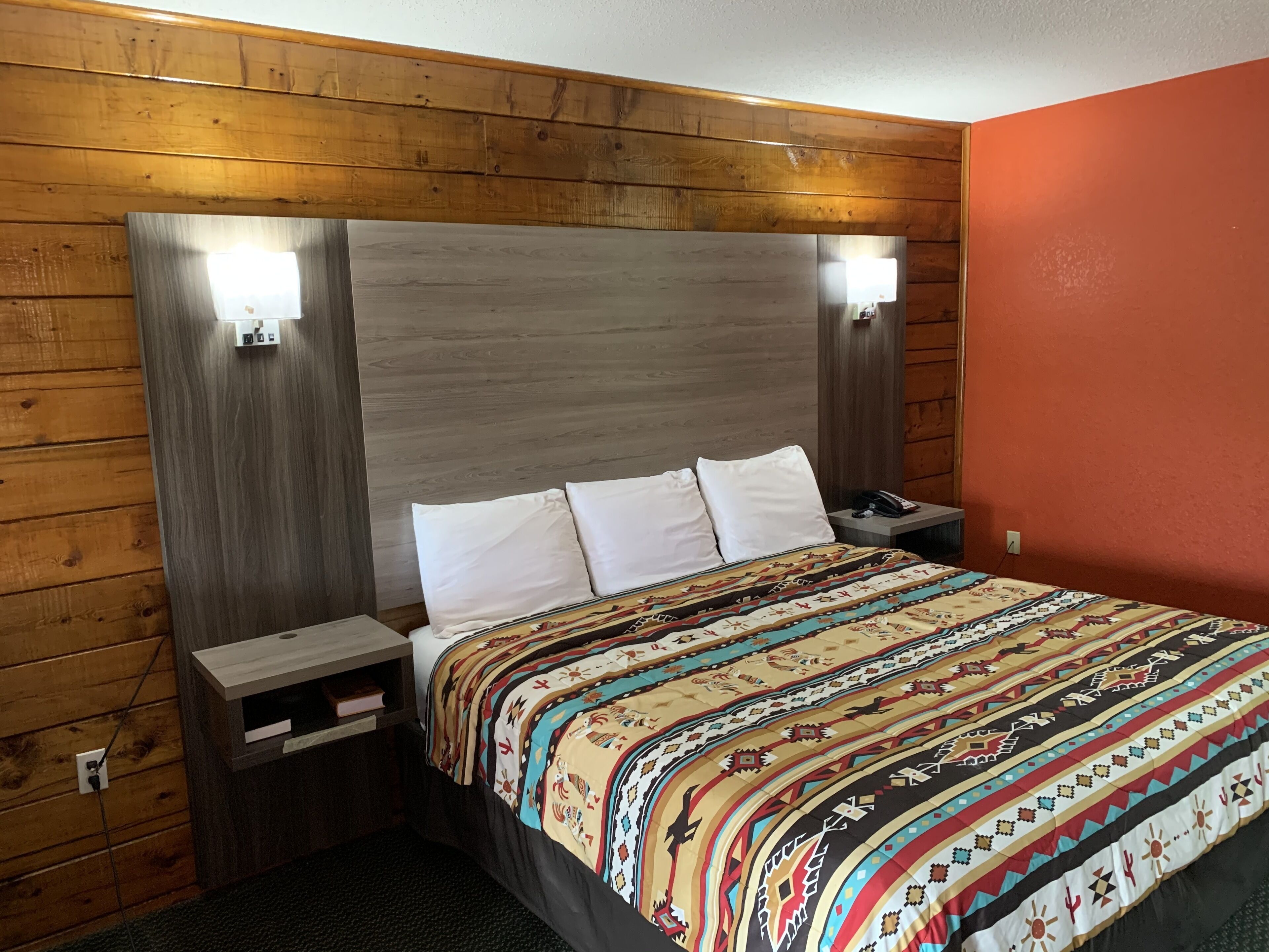 A hotel room with a wooden headboard, a patterned bedspread, and a nightstand with a phone and a lamp.