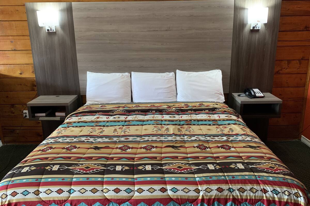 A bed with a patterned bedspread, two white pillows, and a wooden headboard with two wall-mounted lights.