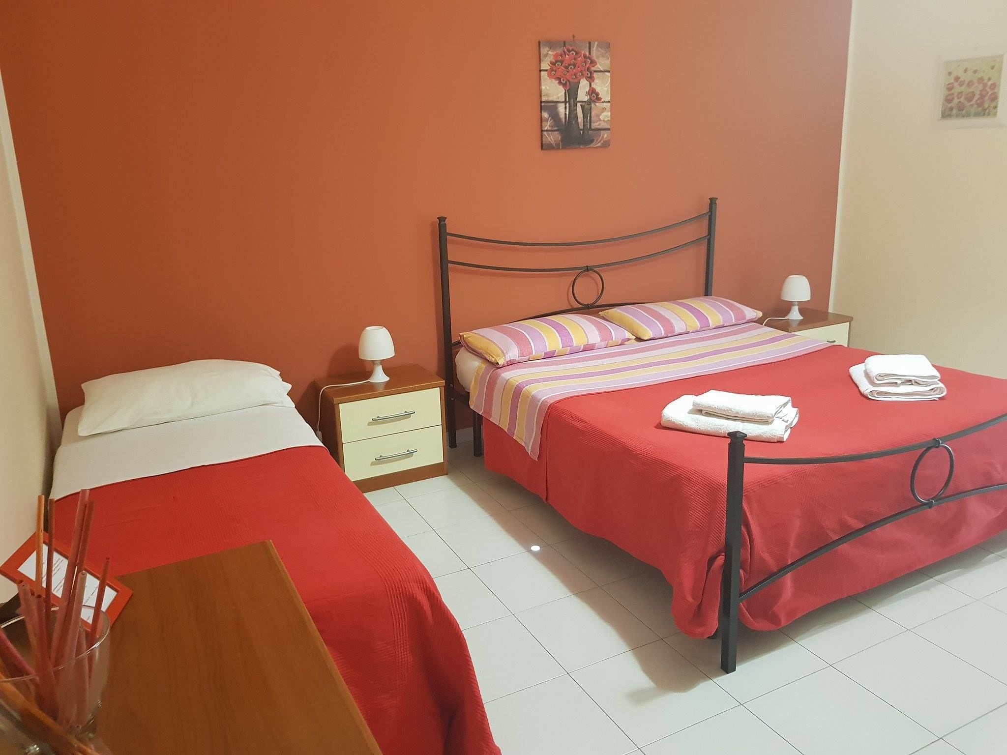 A bedroom with two beds, each with a red bedspread and white pillows. There are bedside tables with lamps, a small painting on the wall, and a wooden chair in the corner.