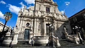 A historic building with ornate architecture, a prominent entrance, and a staircase leading to the main door. - The Artists (Acireale)