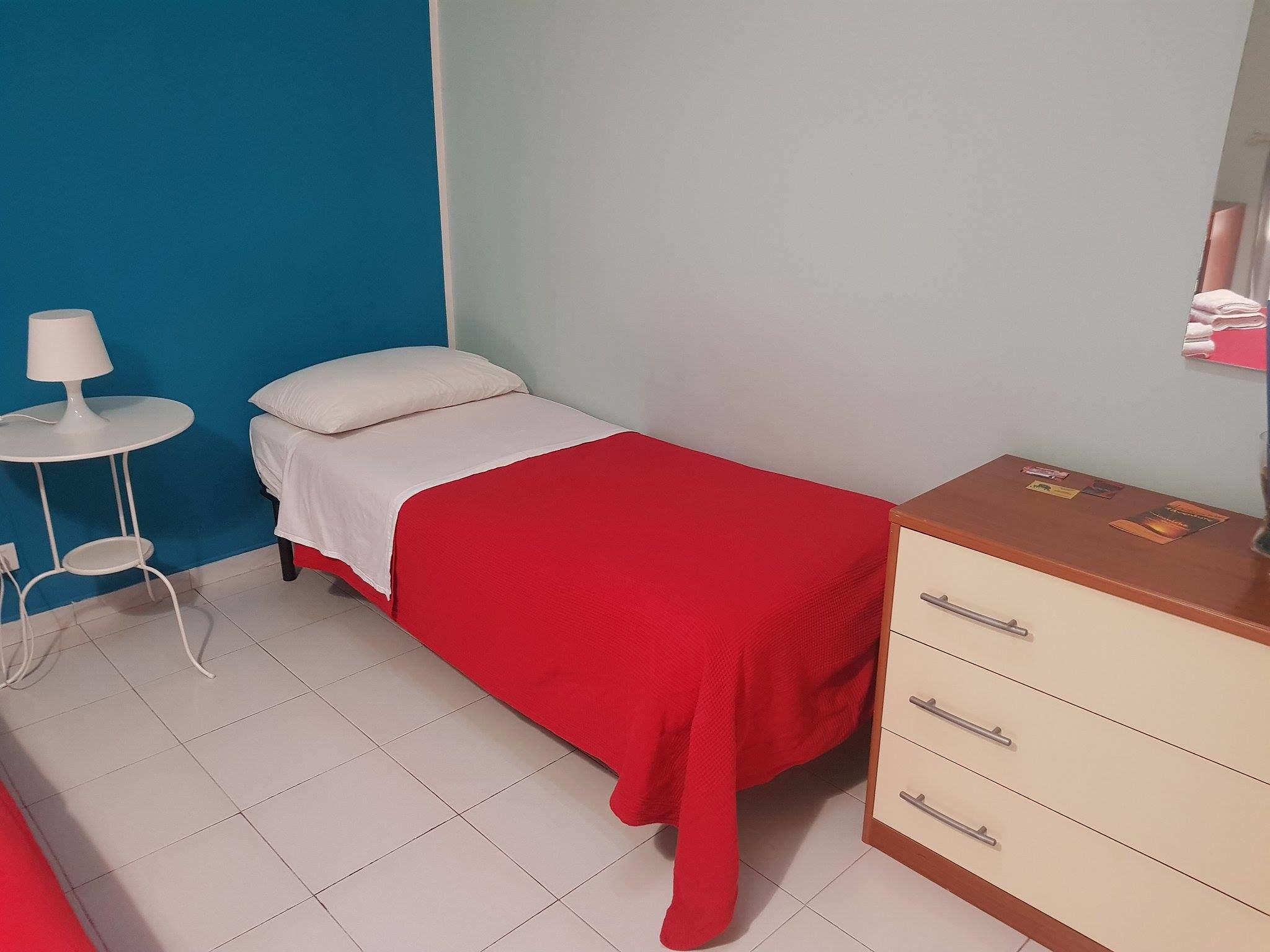 Habitació petita i ordenada amb un llit, una taula de nit, una làmpada i un calaixera.